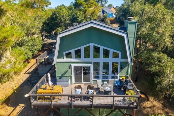 Green barn-style house with large windows and deck surrounded by tall trees, aerial view