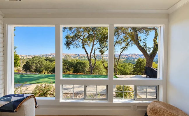 Living room with large windows overlooking green lawn, mature trees, and rolling hills with clear blue sky